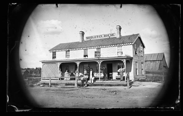 A group posed on the porch of the Middleton House, a two-story frame building with lattice and cut work, located across the intersection from J. Green & Co. Store. A hitching rail is in front of the building as well as a hitching post.