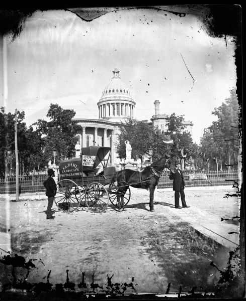 Issued as a stereograph on green mount entitled "The Artist." Two men stand by a wagon on which is written "A.L. Dahl Landscape Photographer." The flag on the side of Dahl's wagon is a souvenir of the Philadelphia Centennial International Exhibition of 1876. A wrought iron fence surrounds the Capitol Park. This Wisconsin State Capitol building, viewed from West Washington Avenue, was erected in 1857 and replaced in 1913.