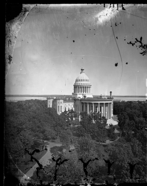 Elevated view of the third Wisconsin Capitol as originally constructed. The trees of the Capitol Park are already so large that they largely obscure any view of the Capitol environs. This photograph was taken by Photographer Andreas Dahl from the roof of the Park Hotel on Carroll Street looking toward Lake Mendota, which is in the background. Interesting details include the striped awnings shading the rooms of the History Society then quartered in the old South Wing, and the white stones that outlined the carriage drive in the park.
