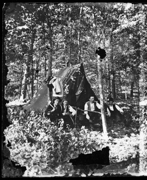 Four men in front of tent in the woods. There is a man with a hatchet next to a clothesline between two trees. This is a Centennial encampment on the shores of Lake Monona.