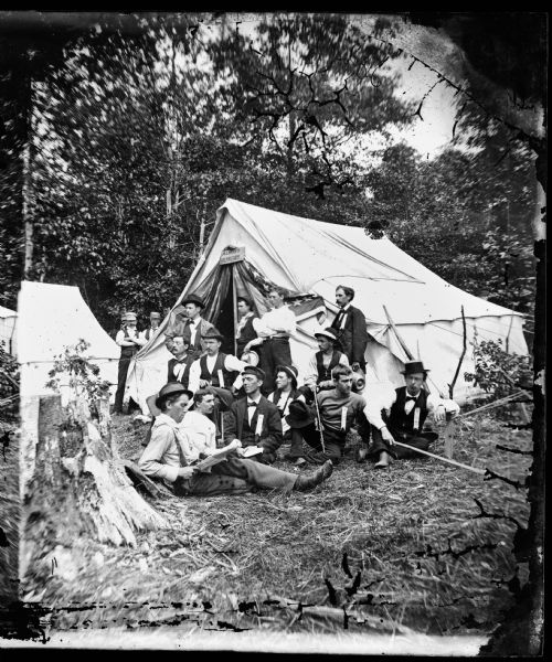 A tent of the Chicago Traveling Club with a sign on it saying "Always Hungry," and men with walking sticks, straw hats and a flute lounging on the ground in front of it. Chicago Traveling Club, Trip #8.