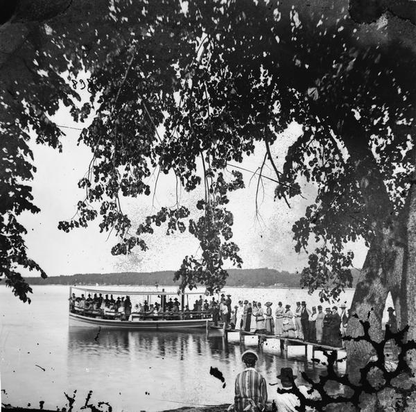 View from shoreline of a large crowd on a pier waiting for a crowded steam excursion boat.