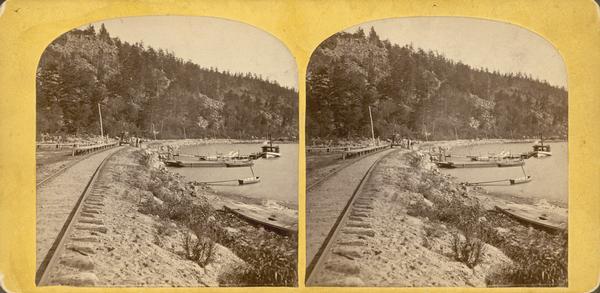 A yellow mount stereograph of the railroad tracks skirting the edge of Devil's Lake, with several boats tied up alongside. From the stereograph series "The Wonders & Beauties of Nature, Devil's Lake, Wis."