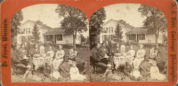 The Reverend Peter Andreas Rasmussen (1829-1898), his wife Ragnhild Holland Rasmussen and their eight children sit around a table in the yard of their house. The table is burdened with large books and a large glass dome containing a floral arrangement. Behind them is an upright and wing frame house.