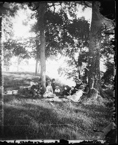 A group, probably at Picnic Point on Lake Mendota in Madison, posed under the trees.  Picnic baskets are behind them.  Mrs. Bashford is the person in Mrs. Ann Fitch's lap.  Next is Mrs. Proudfit.  Mrs. Fitch is lying down.  Dr. Ward, mother, Mr. McConnell, Mrs. McConnell, Mrs. Ward and some Chicago visitors.
