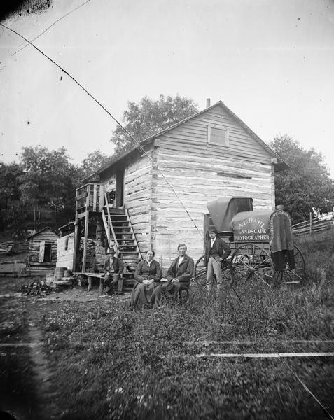 Andrew Dahl's wagon is on the side of a two-story log house in Blue Mounds. On one side of the wagon cover is "A.L. Dahl Landscape Photographer." On the back of the wagon cover is "Stereoscope Views for Sale." Four people, a split-rail fence and other log structures are also in the photograph.