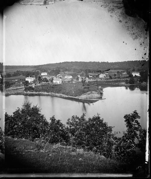 A view of the village from a nearby bluff across a pond. Men, women and children, standing spaced apart, are posing in the field near the pond.