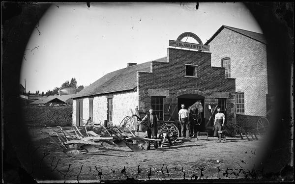Asa Preston (1816-1892) stands in front of his blacksmith's shop with his sons David (1840-1904) and John (1842-1898) behind him and a variety of items made at the shop, particularly plows. The building, no longer standing, was located on Cresent Street.