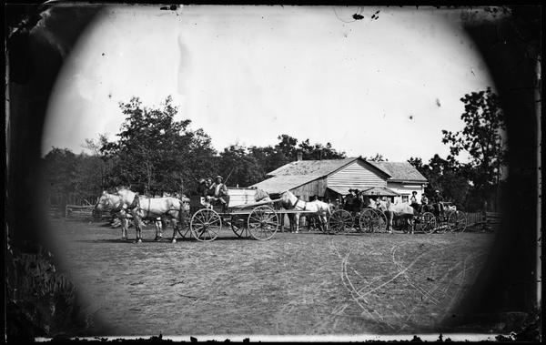 Men in wagons posed in front of a rural post office. They brandish guns, whiskey bottles and possibly a harmonica with humorous intent.