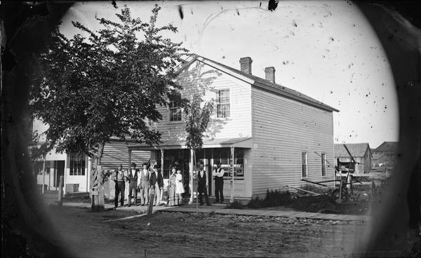 People stand on a sidewalk and porch in front of a frame store. Farm machinery is on the side of the store.
