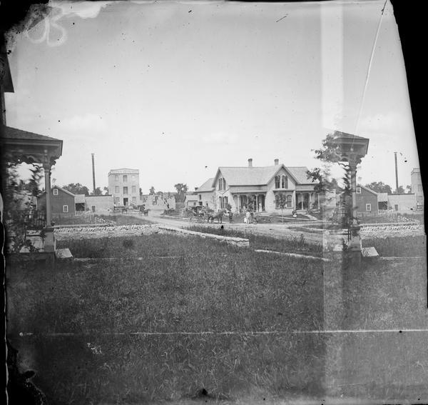 View of Waupun Stone Mills, established in 1849, in the background.  Construction of an addition is underway. People are in the foreground in front of a brick house with a bay window. Another house in the foreground has a stone fence surrounding it. A carriage and wagons are in the street.