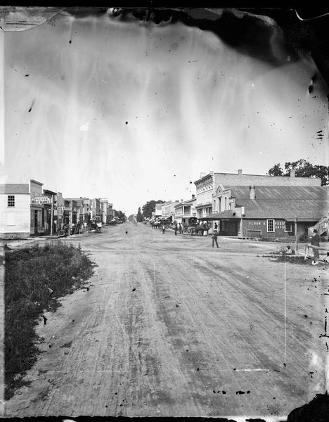 View of a main street looking towards The Fulton Meat Market, P. Carrington Harness shop, a livery stable, feed store, restaurant and bakery.