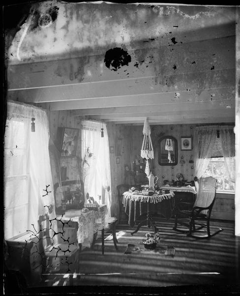 Interior view of the Perry Lutheran Church parsonage, home of the Reverend Jacobson and family. Included in the view is a rocking chair, peacock feathers in a vase, lamps, pillows, tables, china, books and a mirror.