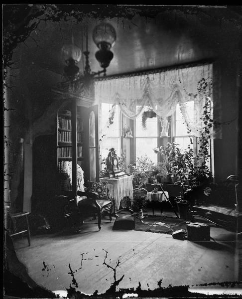 View of a parlor interior (probably that of Herman Amberg Preus's parsonage) with a bay window and draperies, a statue, plants, an ornate clock on a table and a chair in front of an opened bookcase.