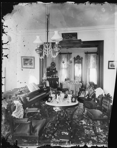 View of two rooms possibly in the Martin K. Dahl home. The room in the foreground has a square entrance way and is filled with a piano, three matched chairs with cloths on their backs and a table in the center with portraits, plants and a statue on it. In the next room are a table and a buffet filled with coffee service pieces, a mirror and windows with lace curtains. (The windows have shutters.) A "Home, Sweet, Home" sign is above the entrance way. Two portraits are hanging on the wall.