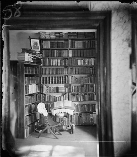 Interior view of Reverend J.A. Ottesen's library, with a chair, a dictionary opened on a stand and a framed photograph on top of books on a shelf. Entitled "Rev. J.A. Ottesen's library," in Dahl's 1877 "Catalogue of Stereoscopic Views."