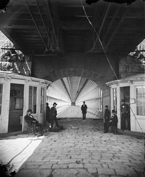 View towards men and women sitting and standing, beneath the lower deck of a suspension bridge (train uses upper deck). There are two booths (for tolls or customs or rail tickets), on the left and right, with Great Western Railway signs in one booth. Stairways to the upper deck are above the both booths. Called "Interior view of Suspension Bridge" in Dahl's 1877 "Catalogue of Stereoscopic Views."