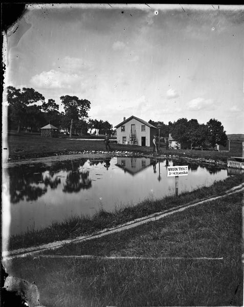 A pond at the Wisconsin State Fish Hatchery (aka Nevin Fish Hatchery, 3911 Fish Hatchery Road) with a sign reading "Brook Trout 3 and 4 years old." Funds for the establishment of the hatchery were received in 1873, and by 1876 it was in full operation. Two men are standing on the opposite side of the pond.