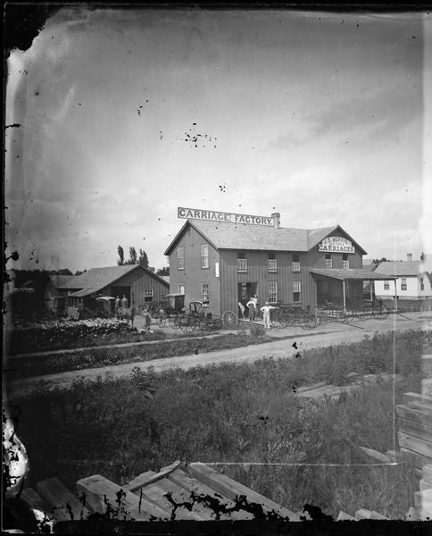 View of the J.S. Morris Carriage and Sleigh Factory, a two-story frame building on Franklin Street in Waupun. Carriages and men in work aprons are in front of the building.