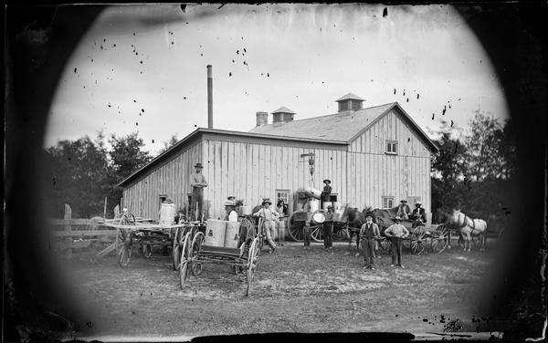 Farmers off-loading milk cans from their wagons at the creamery. One man is operating a hoist, lifting a can to pour the milk down a chute.