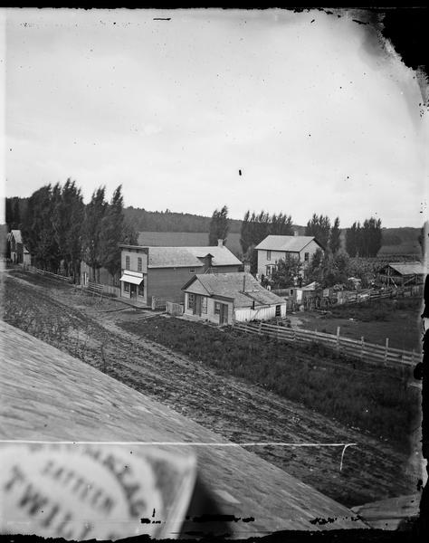 A view from the roof across an unpaved road towards a building across the street from A.L. Dahl's photography studio. The two-story storefront is a general store whose sign reads: "Dry goods, groceries, drugs, hardware, crockery, hats, caps, boots, shoes, farmer's produce." To the right of it is what may be Dahl's brother's livery stable. Behind it is a long board and batten building housing Dahl's studio.