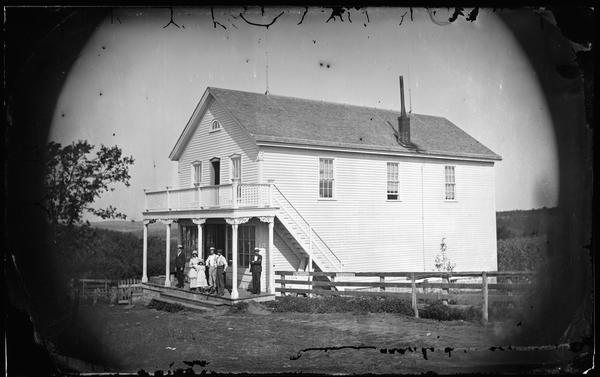 A family standing on the porch of the Onon Bjorn Dahle Store. The store has a second floor balcony across the front, and steps and a fence on the right side. Lightning rods are on the store roof. Two signs on the fence read: "See McCormicks Harvesters!" and "Try and Advance".