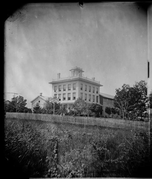 View of the Koshkonong Hotel (later the Lakehouse Inn), a stone and brick hotel with lots of windows, a parapet roof and a small structure on roof, on Maple Beach Road. A man is leaning on a picket fence.