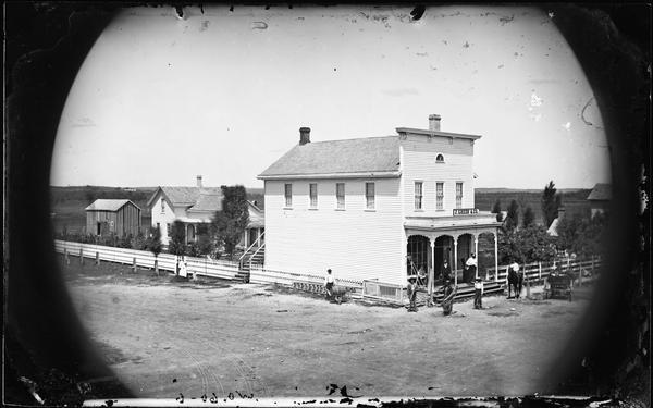 Elevated view of the J. Green & Co. Store, a frame building with a false front, a third-story fan window and two full windows. Men stand nearby with farm implements. A picket fence surrounds the store and other buildings behind it.