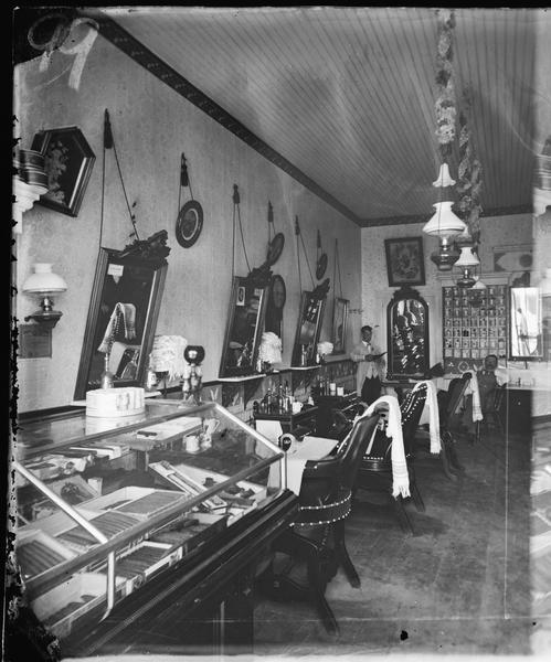 A highly decorated barber shop interior, possibly Wellman's, with a view of chairs, mirrors and a glass display case with cigars. In the back of the shop is a man standing in the corner on the left, and another man sitting on the right.