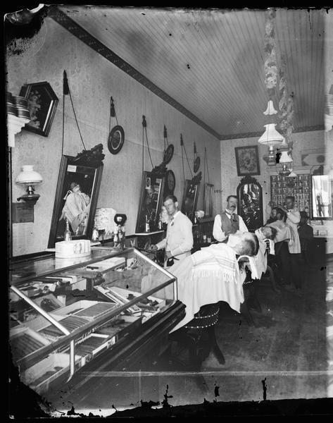 A highly decorated barber shop interior, possibly Wellman's, with three barbers and three customers. Two men are about to get shaves. A glass display case, foreground, includes boxed cigars.