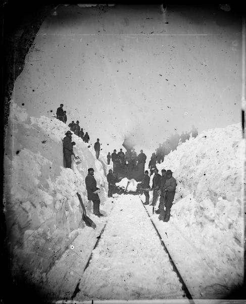 Winter scene with men along railroad tracks in snow and a train engine behind the men. "The Grand and Majestic Snowbanks on the Madison and Portage Railroad, 24th of March, 1875," possibly one of four views of "15 to 30 ft. high snow-banks" in Dahl's 1877 "Catalogue of Stereoscopic Views."
