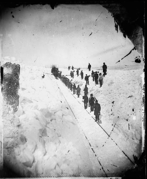 Winter scene with train crew shoveling snow off tracks of Madison-Portage Railroad.  Stereo. "The Grand and Majestic Snowbanks on the Madison and Portage Railroad, 24th of March, 1875," possibly one of four views of "15 to 30 ft. high snow-banks" in Dahl's 1877 "Catalogue of Stereoscopic Views."