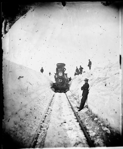 Winter scene with men standing on snow banks near a Madison-Portage Railroad locomotive. "The Grand and Majestic Snowbanks on the Madison and Portage Railroad, 24th of March, 1875," possibly one of four views of "15 to 30 ft. high snow-banks" in Dahl's 1877 "Catalogue of Stereoscopic Views."