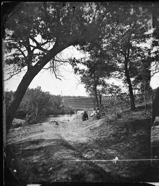 View, from the north bank of the Wisconsin River, of the 1899-foot railroad bridge in Merrimac. Two women and a man are sitting on the riverbank in the foreground.
