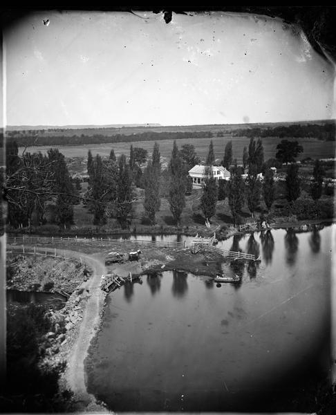 View from above, of a pond and a farmstead.  A man is in a boat on the water and another man is in a wagon on shore near a fence.  In the background are some farm buildings and a two-story frame house with shutters.