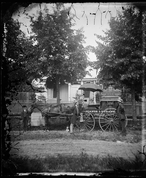 Two men in hats stand by a wagon loaded with an organ. The wagon carries the sign "pianos & organs." A boy holds the horse. Behind them is a picket fence and a frame house with carpenter's trim.