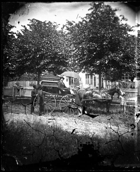 Piano and organ wagon in front of frame house.  Piano and organ man on his wagon with boy leading horse and three men standing near wagon, women in front of a picket fence.  Frame house with shutters closed in bay window, trellis and clothes hanging in yard.