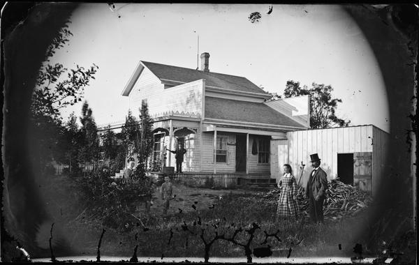 People standing in front of a house with a false storefront. A man is standing on the porch by a hand-pump. There is a woodpile in the yard behind a woman and a man on the right.
