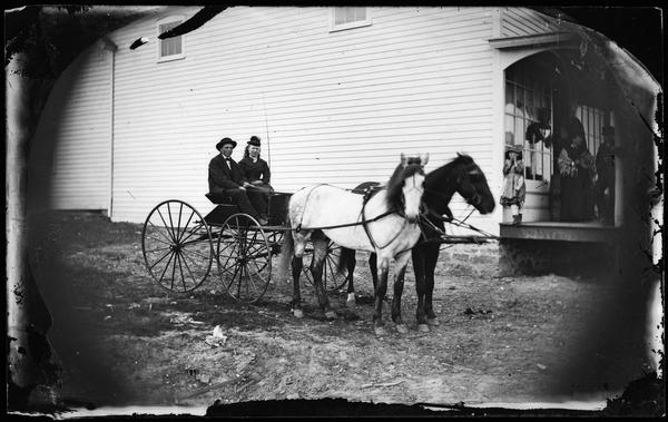 A couple sits in a horse-drawn wagon.  In the background are a girl and another couple on the porch of a commercial frame structure, probably a store.