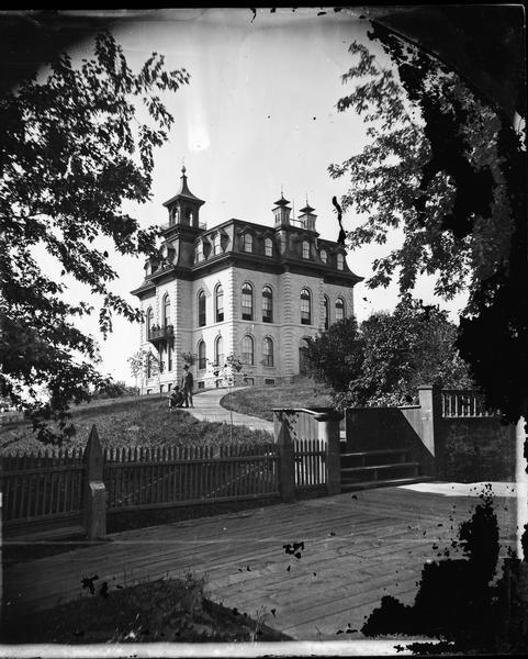 View across wooden platform of a brick high school on a hill surrounded by a fence. The school has a mansard roof and what appears to be a bell tower above the entrance. There are a two students posing on the path leading up to the school, and a few more students are on the balcony of the school.