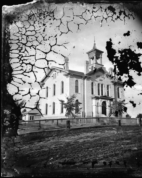 Two-story brick building built in 1870 with arched windows, corner brackets, bell tower with elaborate cut work, four chimneys and fence in front. Inscription above the door reads "Public School" and "Erected A.D. 1870."