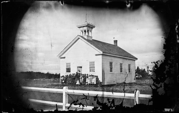 One story schoolhouse with bell tower on roof and group of students and teacher in front. The scho0l is in a rural setting with a fence in the foreground.