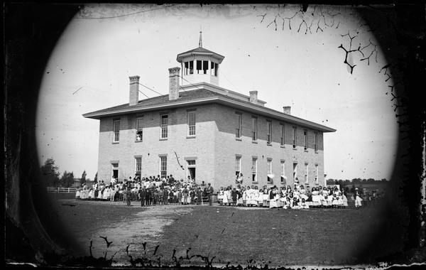 The Mazomanie School, later the high school. A large group of students and teachers are standing in front of the two-story brick building with octagonal cupola, and perhaps a bell. Three students are looking out from the front second-story window, and two students from a lower side window.