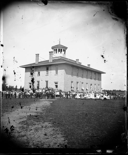 The Mazomanie School, later the high school.  A large group of students and teachers stand in front of the two-story brick building with octagonal cupola and perhaps a bell.  Three students look out from the front second story window and two students from a lower side window.