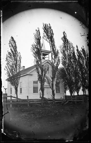 A one room Greek Revival schoolhouse with bell tower. A boy is in front, wearing a broad-brimmed straw hat and climbing a tree.