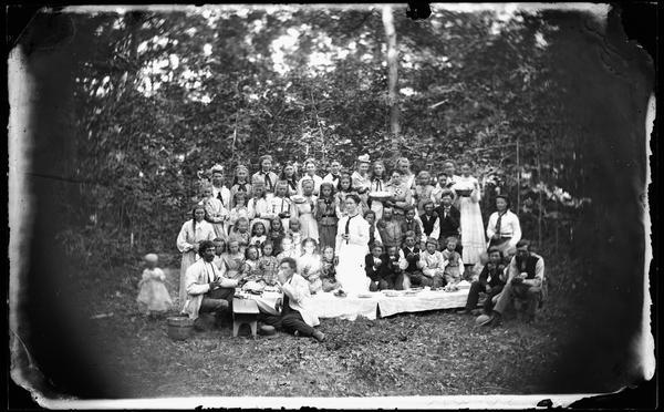Mixed group, mostly children, having a picnic in the woods, perhaps connected with the dedication of the East Blue Mounds Lutheran Church.