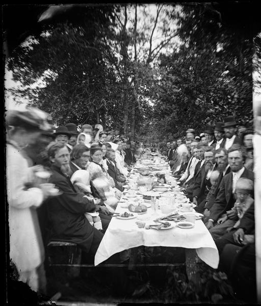 "View of the Dining Table" from the "Dedication of Blue Mounds Church" section of Dahl's 1877 "Catalogue of Stereoscopic Views", showing a long banquet table outdoors, with men, women and children sitting and standing along the sides. The congregation of the East Blue Mounds Lutheran Church and visiting ministers were gathered for the church dedication August 30, 1876. The church was formerly Norsk Evangelisk Kirke, built in 1868.