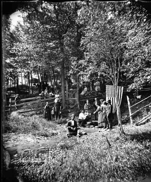 View towards a large group of people with a U.S. Flag hanging from a tree. The group is gathered around a stream near a wood fence and gate under trees. There is a log structure in the background. Perhaps a Fourth of July picnic.