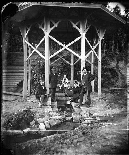 "Mineral spring pavilion with visitors taking the waters." Seven adults, including a man in a top hat, five children and a woman are posing in a gazebo built over the Iodo-Magnesian Springs northwest of Beloit in preparation to drinking the medicinal waters.