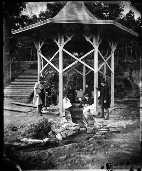 "Mineral spring pavilion with visitors taking the waters." Well-dressed men, women and children are posed in a gazebo built over the Iodo-Magnesian Springs northwest of Beloit in preparation to drinking the medicinal waters. The springs were developed by Thomas Crowder Chamberlain and his brother in 1875. Eventually they were given to Beloit College.